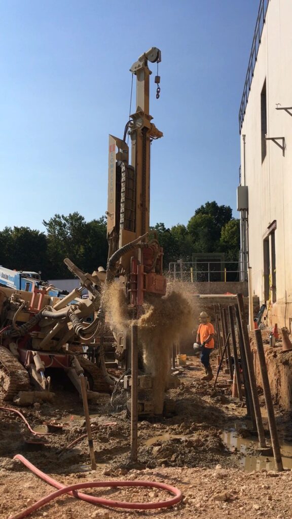 A construction worker wearing an orange vest and hard hat operates heavy machinery on a construction site. The machine drills into the ground, sending soil into the air. A white building is partially visible in the background with trees behind it.