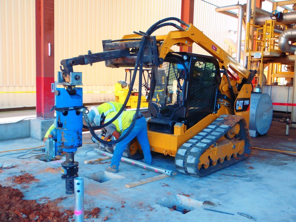 Workers operate a yellow skid steer loader with a blue drilling attachment, working on a concrete surface. They wear safety gear, including helmets and reflective vests. The background features industrial pipes and structures.