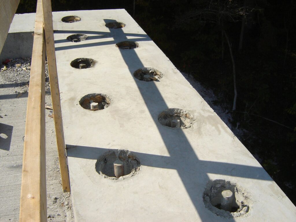 A concrete slab with several circular holes, each containing a protruding metal bolt. Sunlight casts shadows of a railing across the surface. The slab is part of an elevated structure, showing a wooded area below.