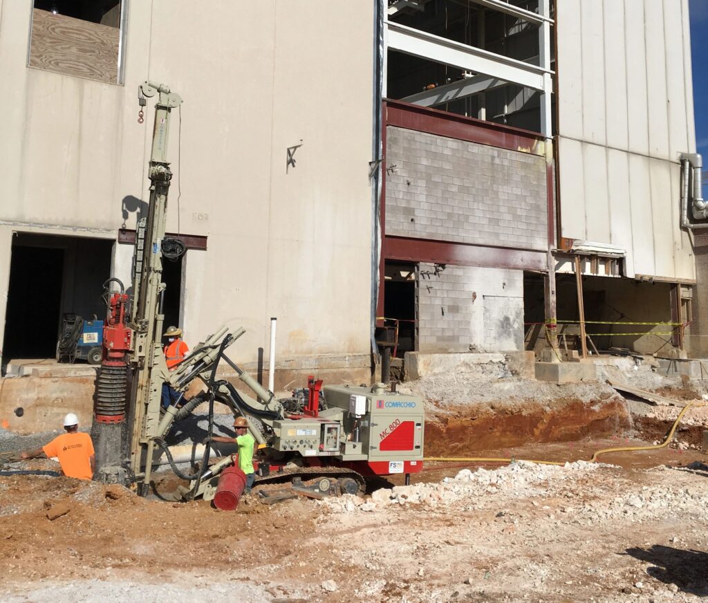 Construction workers operate drilling equipment at an industrial site. They wear safety gear and work near a large building with exposed sections. The ground is partially excavated, revealing layers of soil and rock.