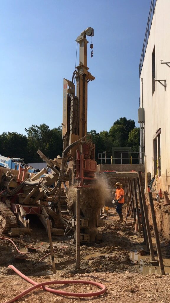 Construction worker operates a drilling machine next to a building. The machine is drilling into the ground, with debris scattering. The worker wears an orange safety vest and helmet. Scaffolding and equipment surround the scene under a clear blue sky.