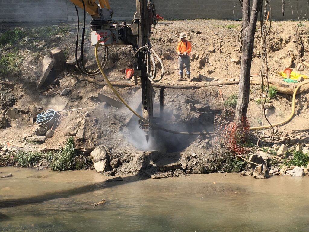 A construction worker in an orange vest and hard hat observes a large drilling machine operating by a stream. Dust and debris surround the drill, and various tools and hoses are scattered on the rocky ground.