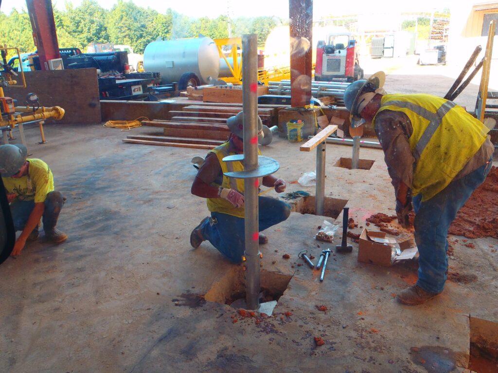 Construction workers in safety gear are installing equipment at a job site. One worker is kneeling and positioning a metal pole into a square hole in the ground, while another worker assists. Various tools and materials are scattered around the area.