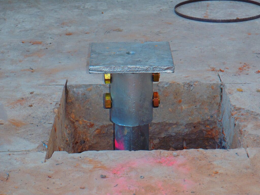 A metal post with a square base set in a concrete floor. The post is secured with bolts and sits in a square hole in the ground. The surrounding concrete has some debris and markings.