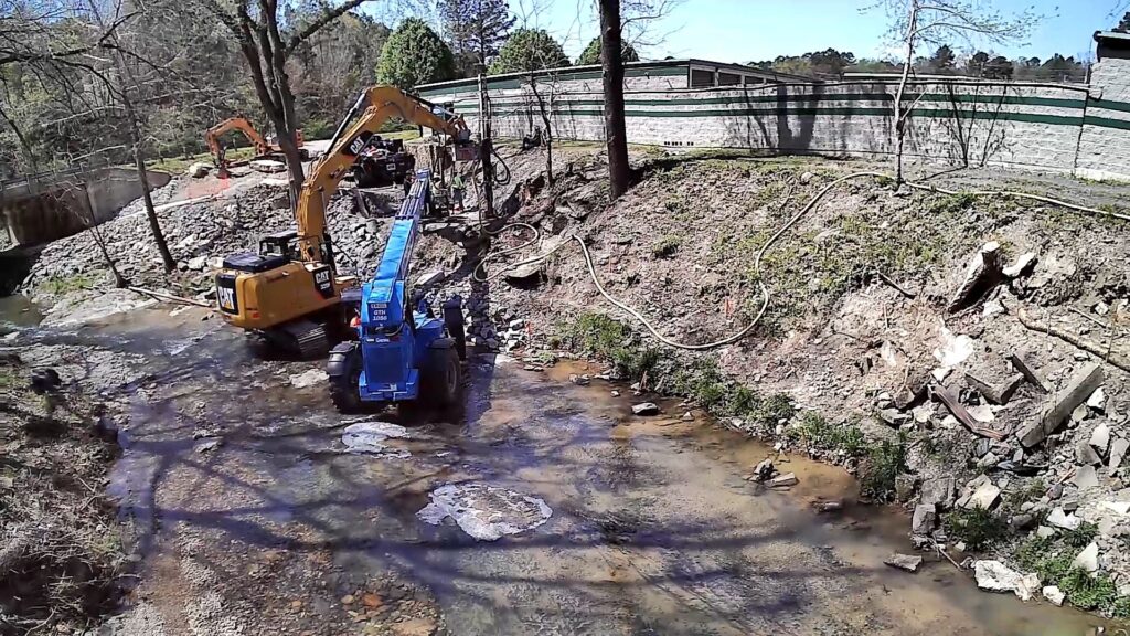 Two excavators, one yellow and one blue, work on a small stream. The area is surrounded by sparse trees and a wooden fence in the background. The ground is uneven with patches of grass and dirt.