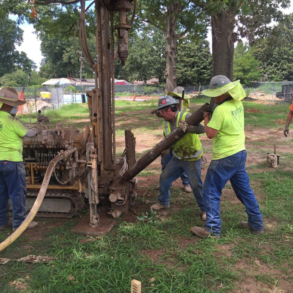 Construction workers operating drilling equipment on a grassy field. Wearing safety gear and high-visibility shirts, they collaborate to manage a large drill, with trees and fenced area in the background.