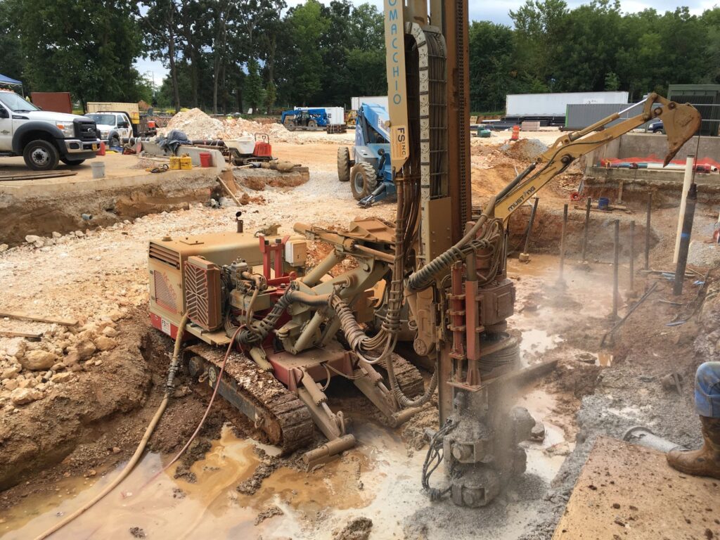 A construction site with a drilling machine operating on the ground. A worker stands nearby, monitoring the process. The site is surrounded by trucks, piles of earth, and construction materials. Trees are visible in the background.