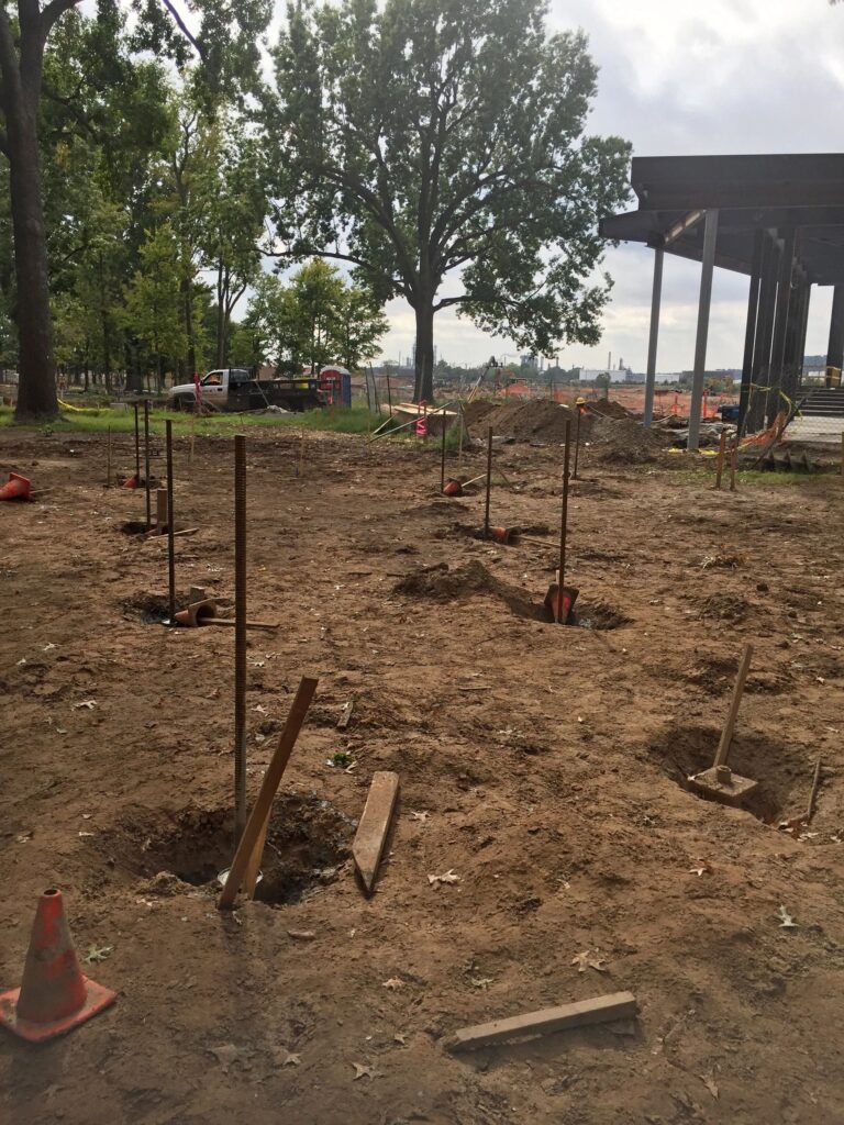 A construction site with several dug holes and stakes in the ground. A few orange cones are scattered around. Trees and a partially built structure are visible in the background, with overcast skies above.