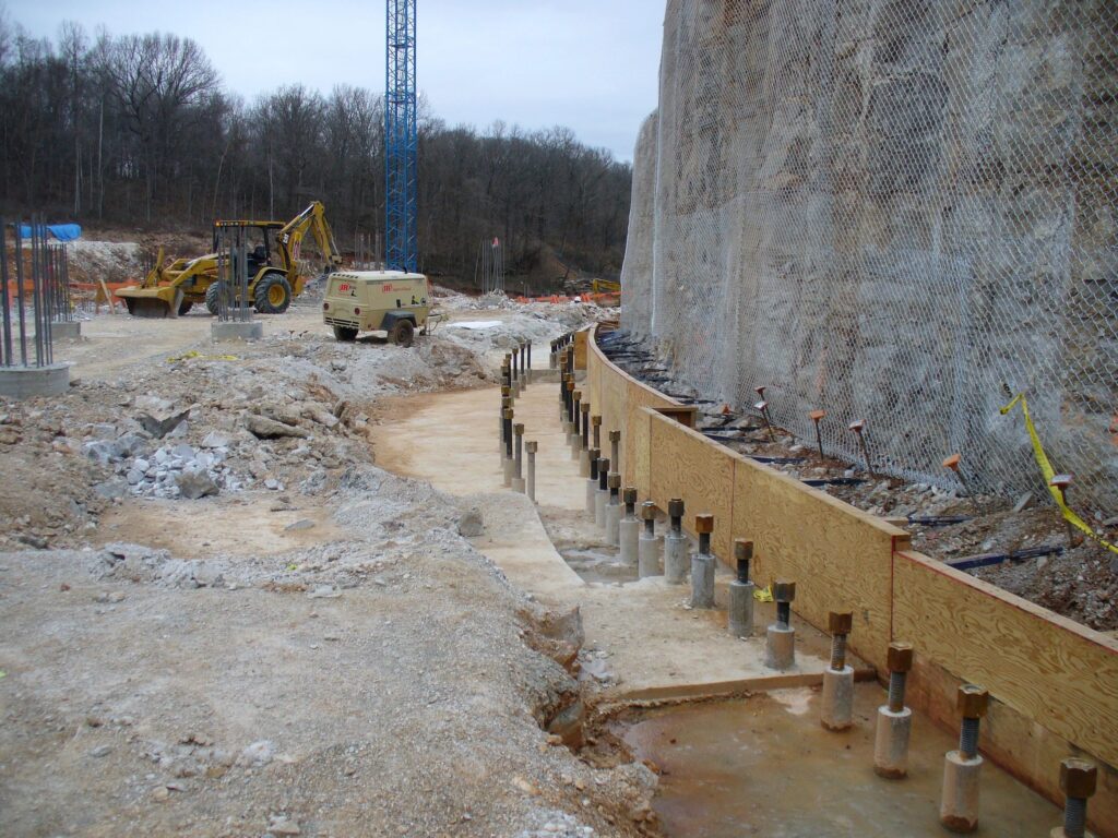 Construction site featuring a large retaining wall under construction with scaffolding and netting. Heavy machinery, including a bulldozer and a cement truck, are present. The area is surrounded by trees and appears to be in a rural setting.