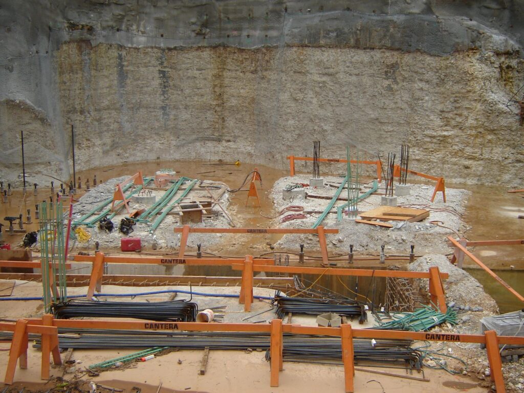 Construction site featuring a large excavation with steel reinforcements and scattered materials. Orange barricades display "CANTERA," and the ground is muddy with puddles. Vertical rods and wooden planks are visible throughout the site.