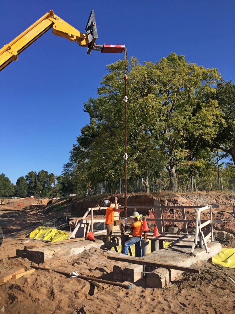 Construction workers operate machinery at a site, using a crane to lift a pole. They are wearing safety gear, including helmets and orange vests. The site is surrounded by trees under a clear blue sky.