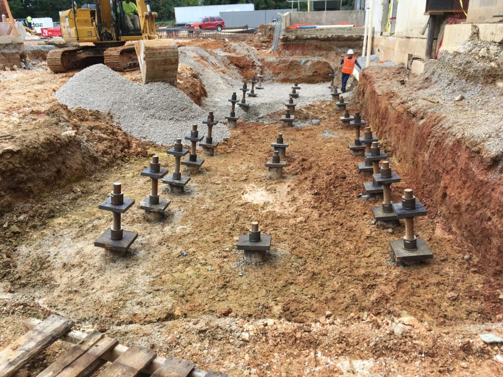 A construction site with several helical piles installed in the ground. A worker in an orange safety vest stands nearby, and a yellow excavator is visible in the background. Soil is excavated around the piles.