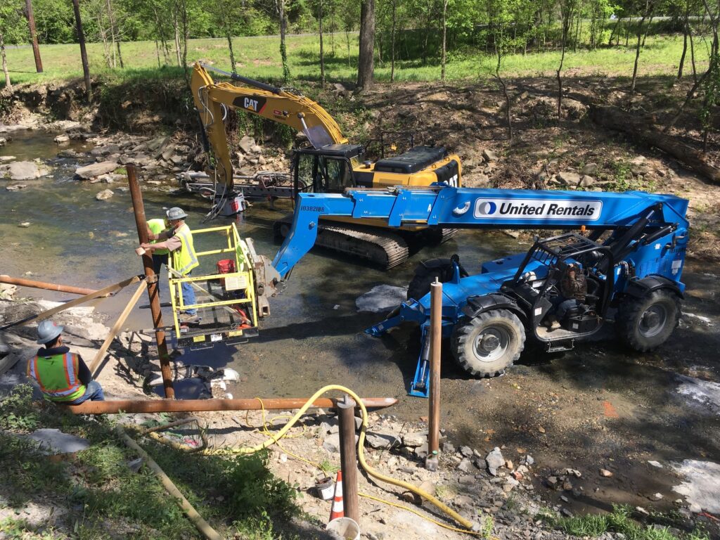 Construction workers operate machinery in a creek area. A yellow excavator and a blue forklift, marked "United Rentals," assist in the work. Workers on a platform manage equipment amidst trees and a rocky, grassy landscape.