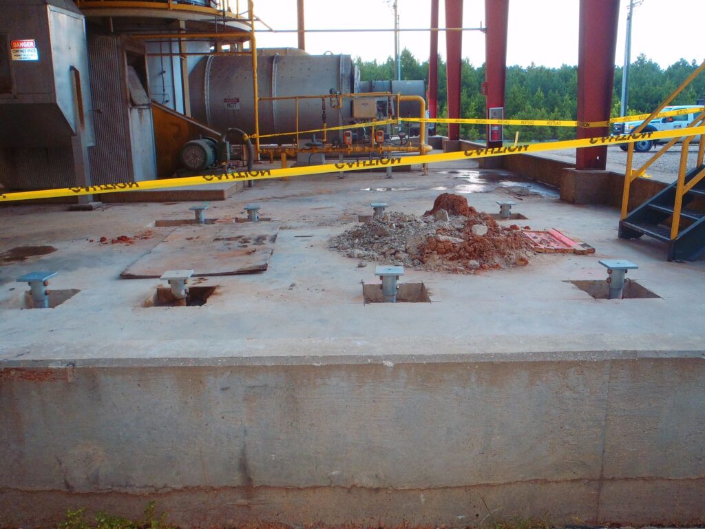 A construction site with a concrete slab and several metal brackets protruding from it. A pile of dirt is in the center of the slab, and yellow caution tape surrounds the area. Trees are visible in the background.