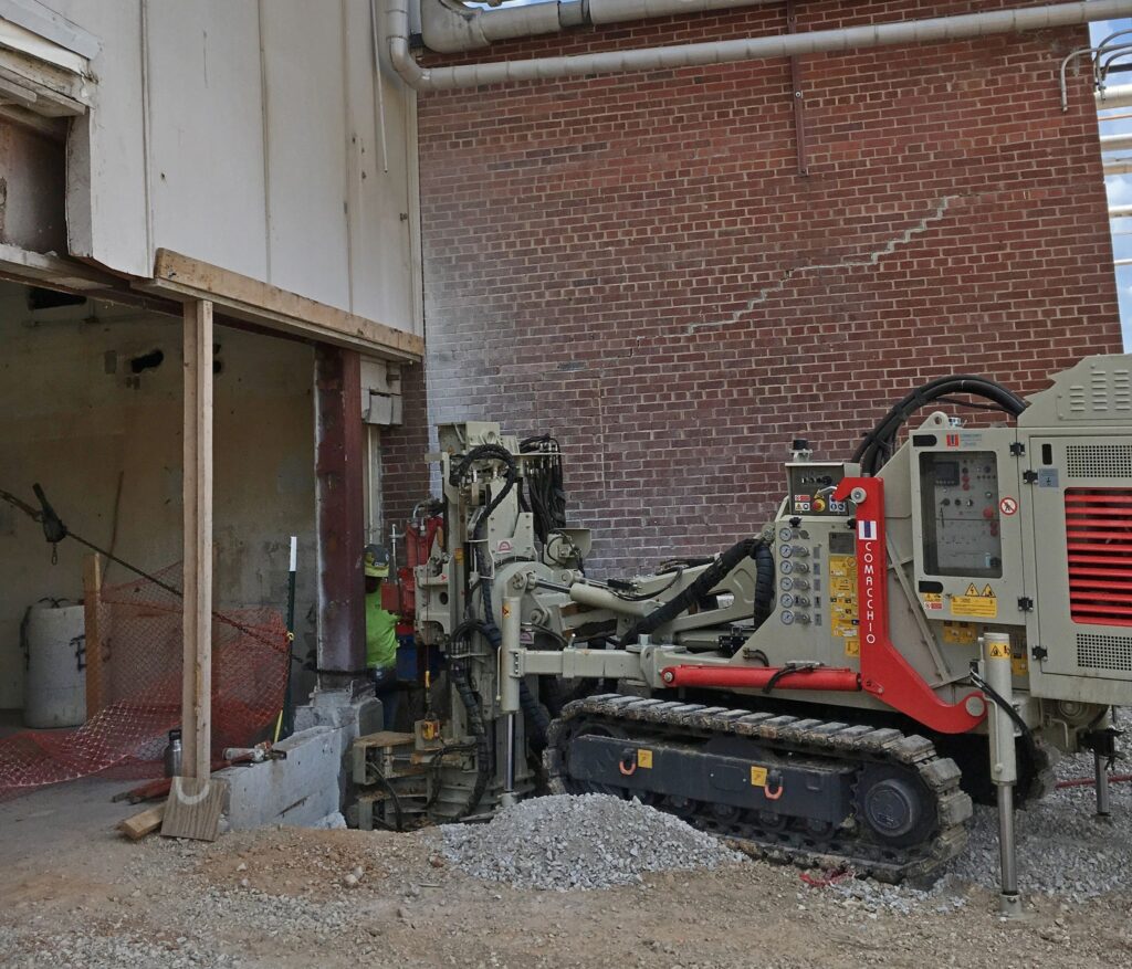 A heavy construction vehicle with tracks is positioned inside a partially demolished building. The room is enclosed by brick walls, with exposed pipes above. The floor is covered with gravel and debris, and a worker is visible near the machine.