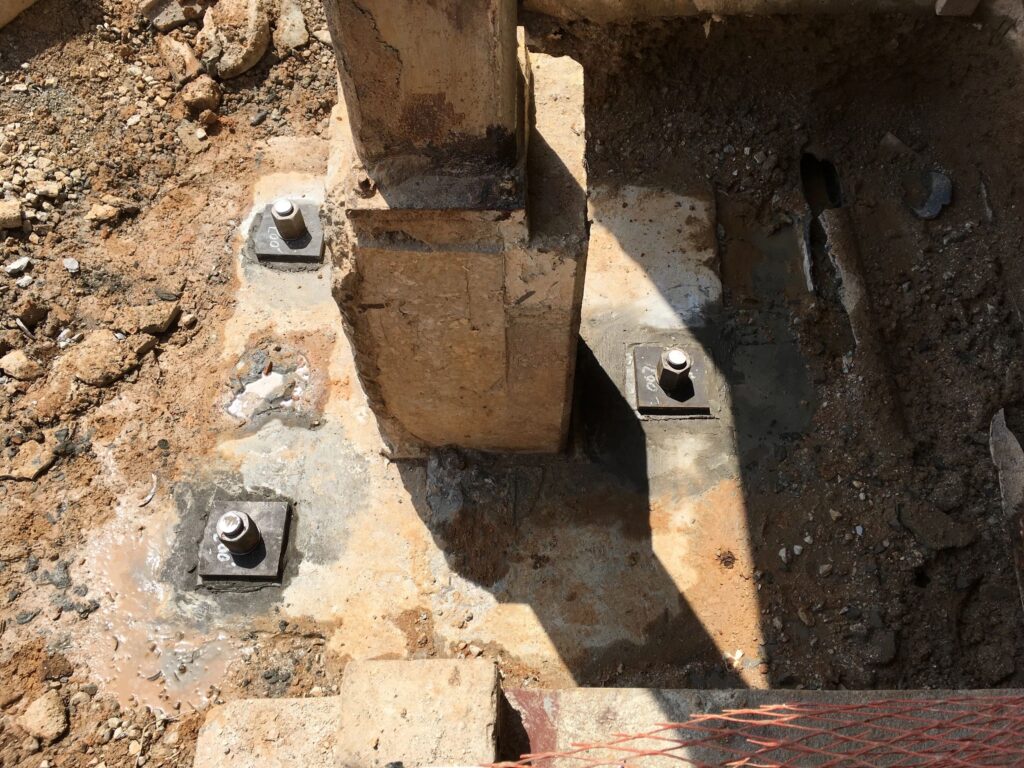 Construction site showing a concrete column with four metal bolts at its base, surrounded by dirt and some debris. The area is partially shaded and features a safety net in the foreground.