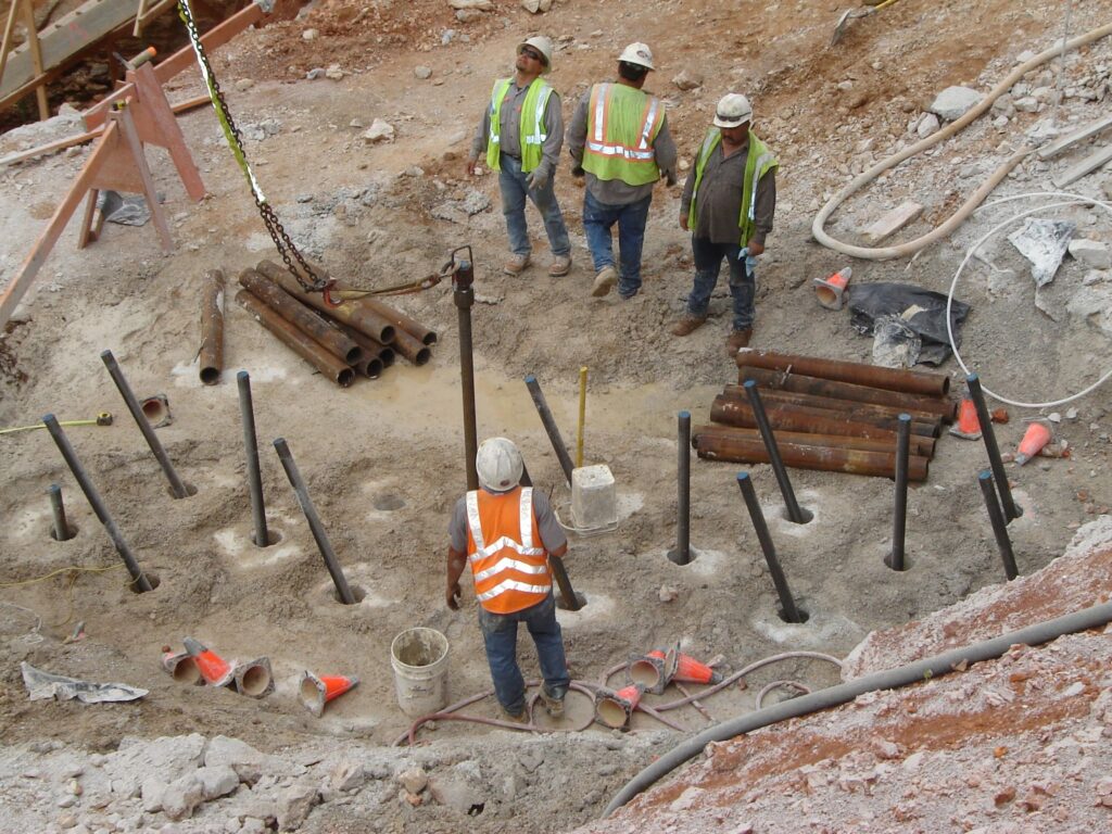 Construction workers in safety gear stand around a drill site at a construction area. Several metal pipes protrude from the ground, and various construction materials are scattered nearby.