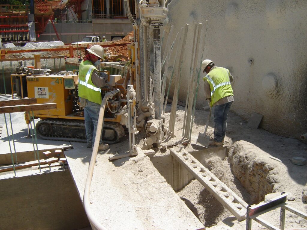 Two construction workers in high-visibility vests and helmets operate machinery at a construction site. They are drilling into the ground, with pipes and cables visible around them. A large concrete wall and building framework are in the background.