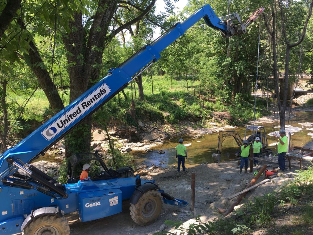 A blue United Rentals crane is positioned near a stream in a forested area. Four workers in safety vests and helmets are placing beams into the ground. Trees and greenery surround the construction site.