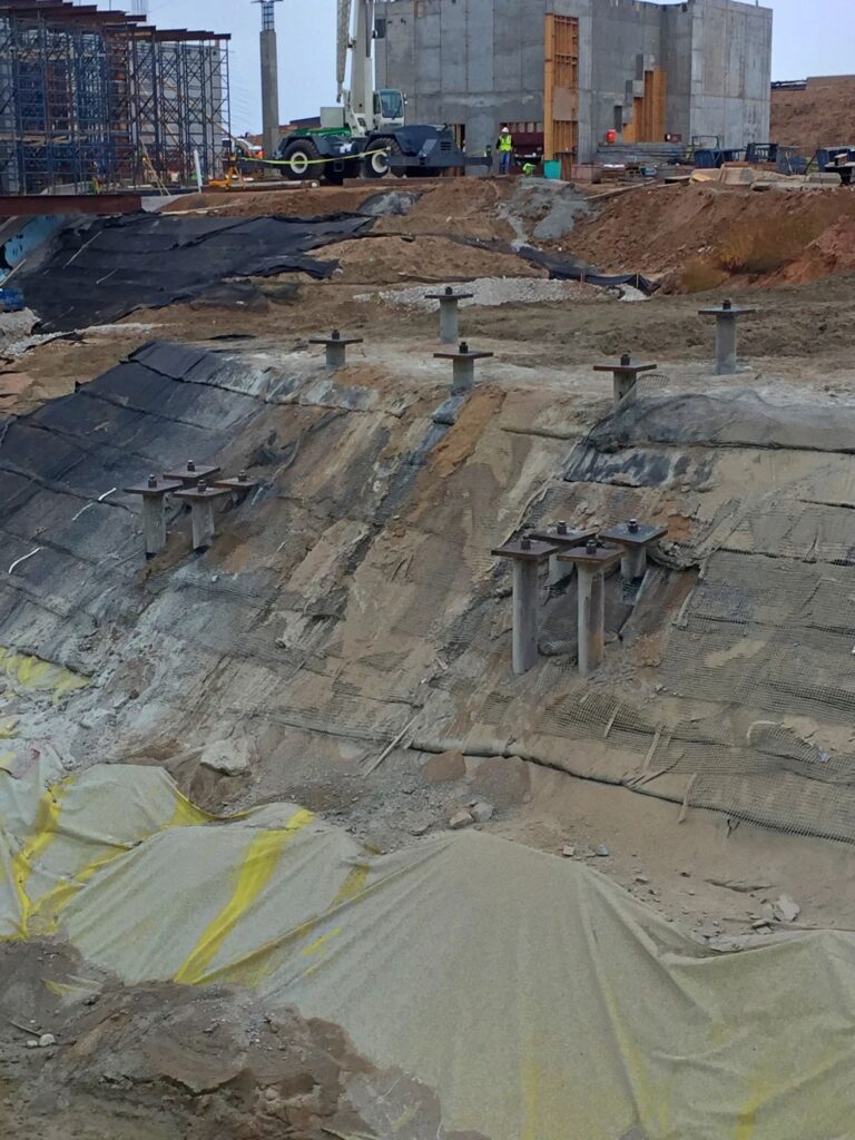 Construction site with concrete structures and steel supports on a sloped terrain. Workers and equipment are visible in the background. The area is covered with geotextile fabric and some sections have exposed soil and gravel.