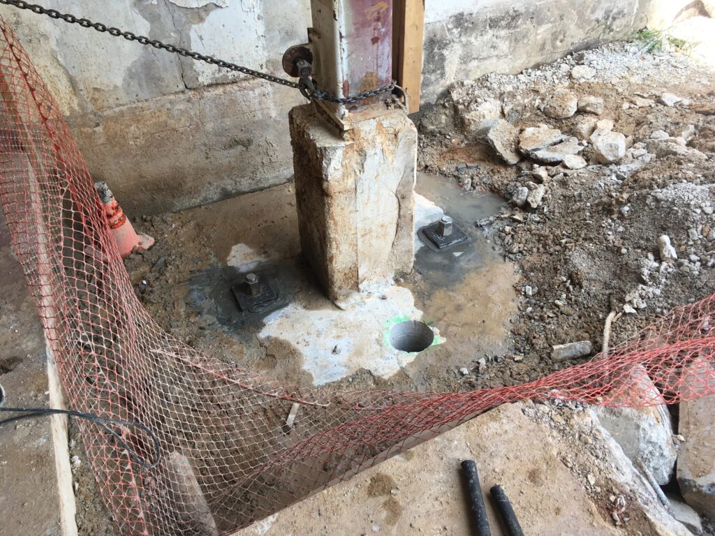 A construction site with a large support post secured to a concrete base. Red safety netting surrounds the area, and there are piles of rubble and loose dirt nearby. Two metal rods are seen on the ground.