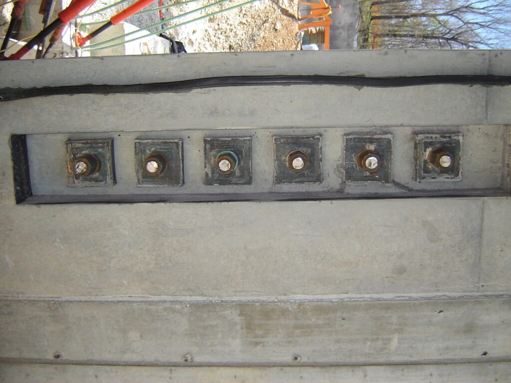 Close-up of six anchored bolts set in a concrete slab, each surrounded by a square metal plate. The bolts are evenly spaced in a row, part of a construction project. A partially visible tree and rocks can be seen in the background.