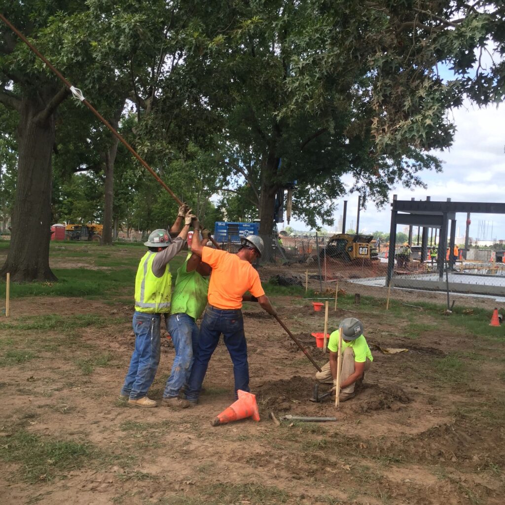 Construction workers are using a long rod to test the ground at a worksite. One worker is in a hole while others assist. They are wearing safety gear and surrounded by trees and construction equipment.