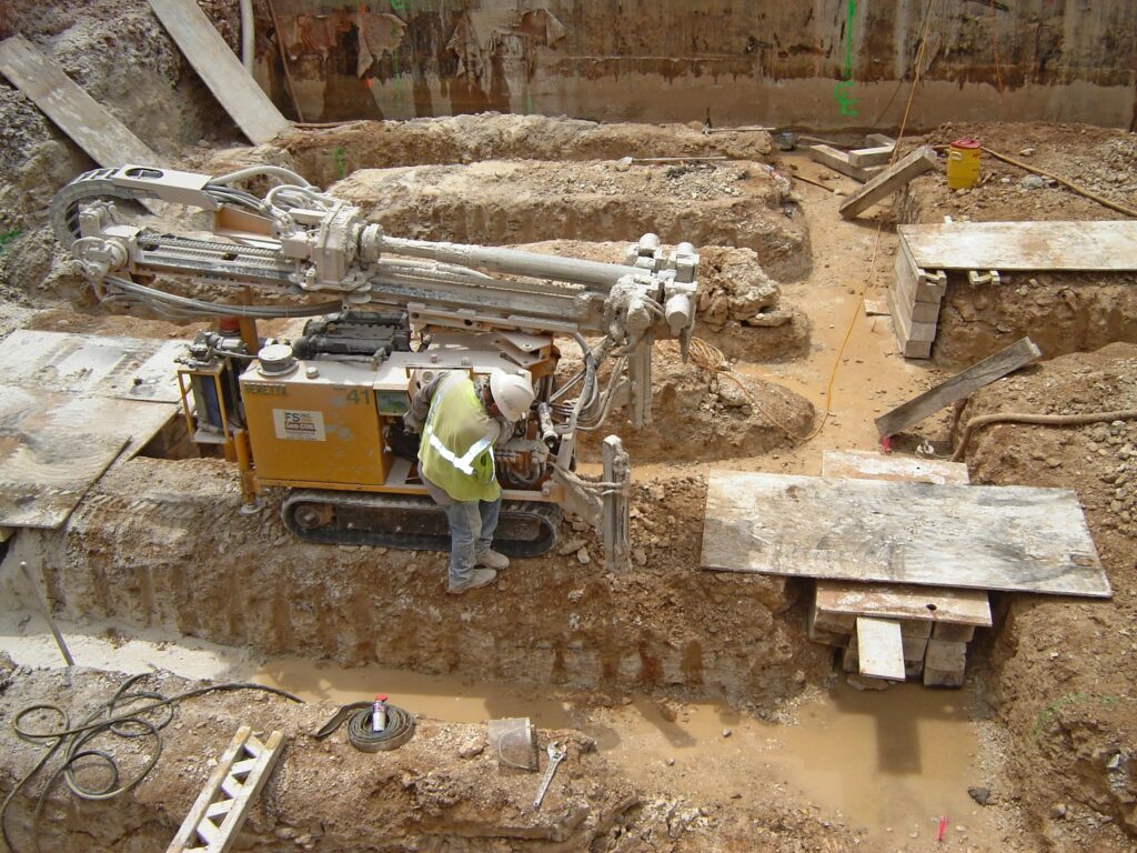 A construction worker operates heavy drilling machinery at a muddy construction site. The ground is uneven with trenches and wooden planks scattered around. The worker wears a hard hat and high-visibility vest.