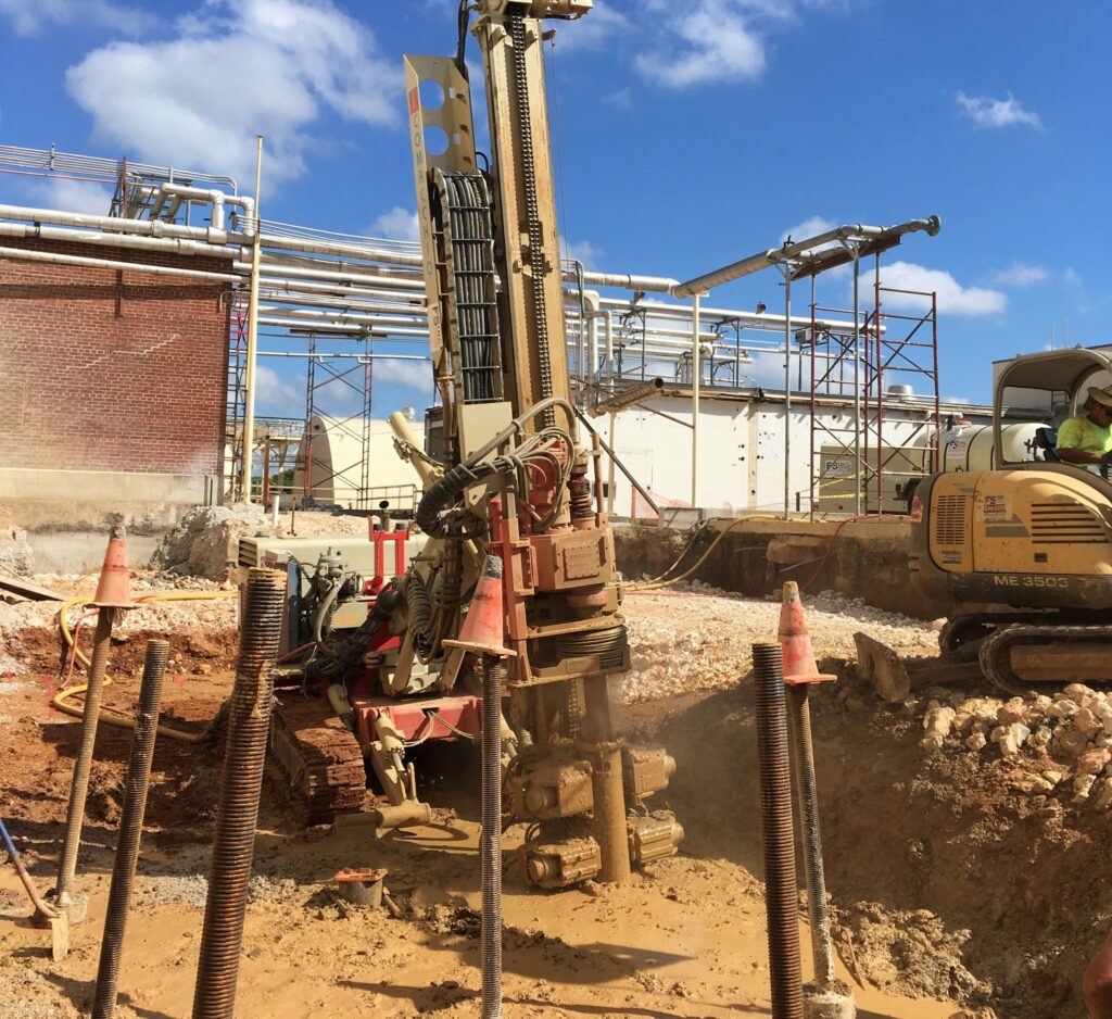 A construction site with a large drilling machine operating in front of a brick building. Several metal rods and traffic cones are visible in the dirt. A worker operates an excavator in the background. Scaffolding and pipes are visible under a blue sky.