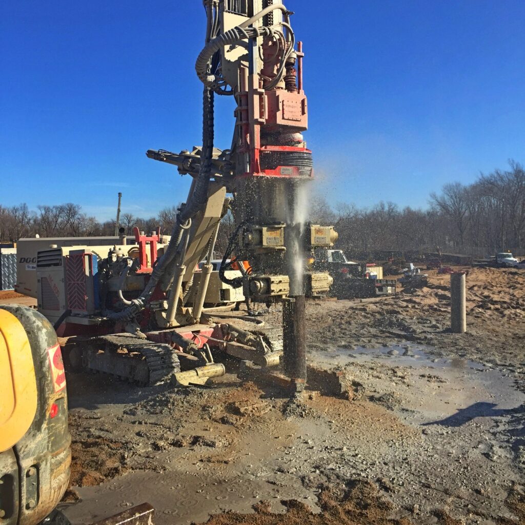 A large industrial drill operates at a construction site, drilling into the ground with water spraying. Heavy machinery and vehicles are visible in the background, with a clear blue sky and bare trees lining the horizon.