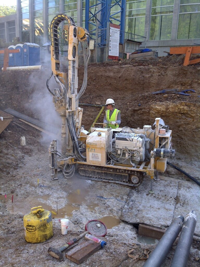 A construction worker operates a drilling rig at a construction site, surrounded by dirt and machinery. The worker wears a safety helmet and vest. Portable toilets and building materials are visible in the background.