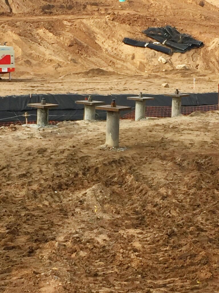 Construction site with several concrete piles emerging from the sandy ground. A black tarp and construction materials are visible in the background. Earthmoving is evident, with uneven terrain and exposed soil.