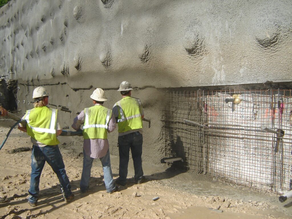 Construction workers in yellow safety vests and helmets operate machinery to apply concrete to a rebar structure on a textured wall. The ground is muddy, and the area appears to be a construction site for infrastructure or a building.