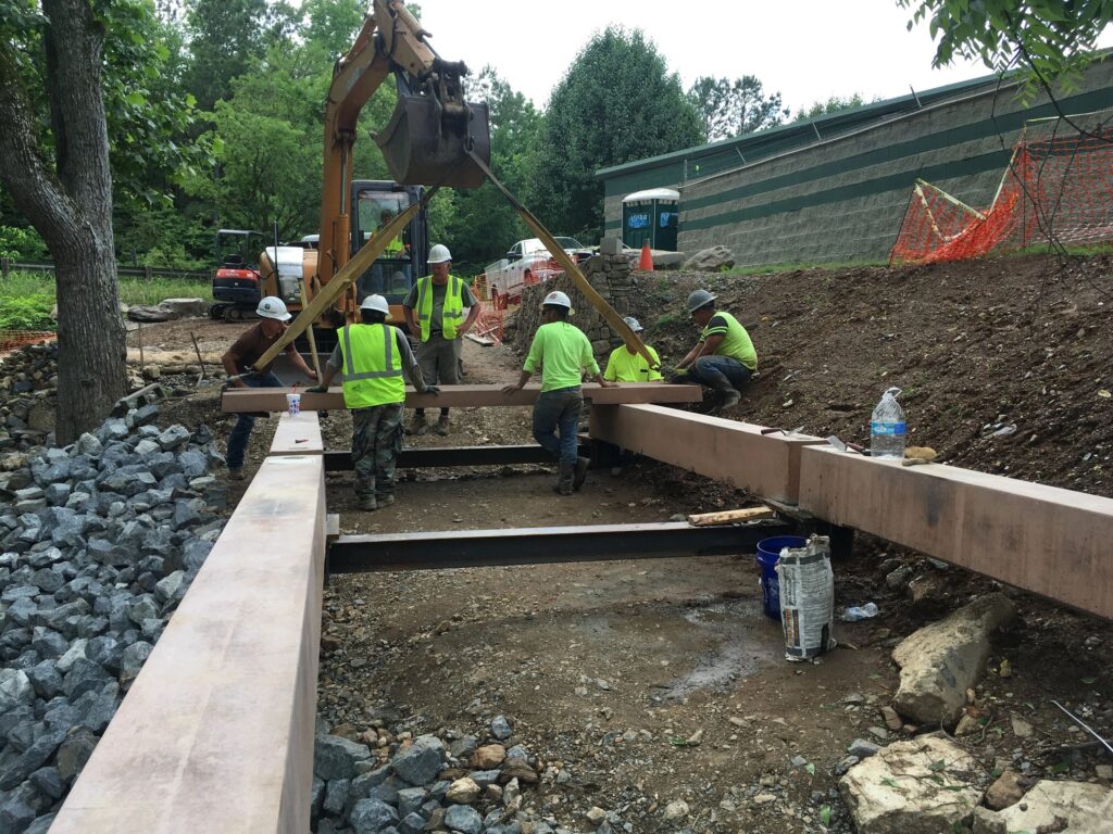 Construction workers in safety gear are positioning large steel beams on a construction site. An excavator helps move the beams. The area is surrounded by trees, gravel, and caution fencing.