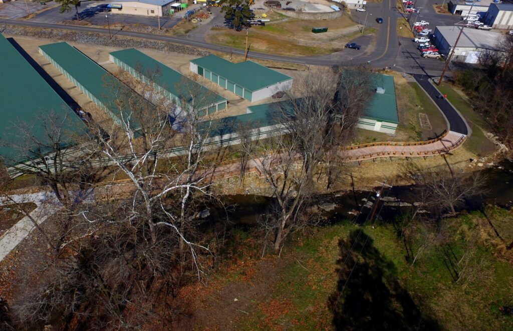 Aerial view of a green-roofed industrial area near a walkway. A row of storage units is visible, with a path running beside a small creek surrounded by leafless trees. Nearby roads and parked vehicles are also seen.