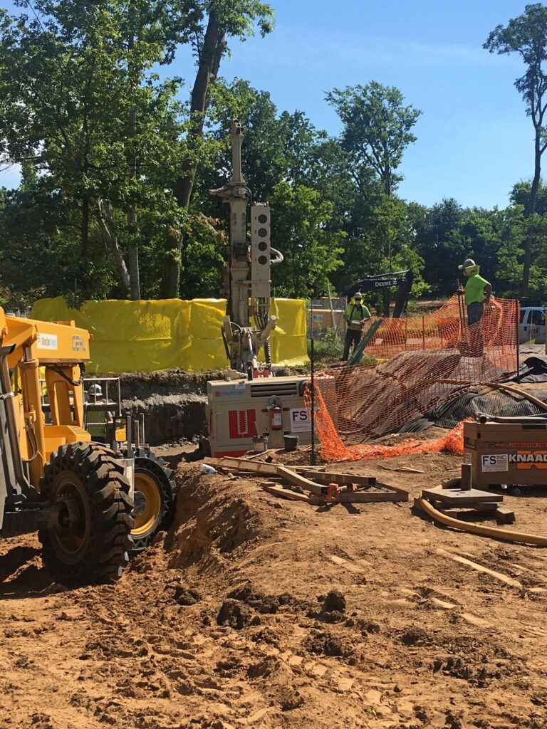 Construction site with workers in green safety vests operating machinery and tools. An excavator and a drill are visible. Yellow safety barriers and orange netting surround the area. Trees and a clear blue sky are in the background.