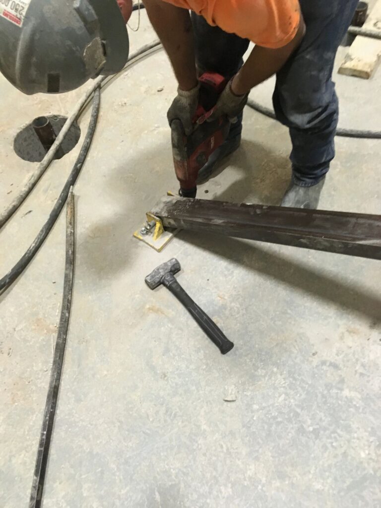 A construction worker in an orange shirt uses a power drill on a metal beam on a concrete floor. A hammer and several cables are scattered nearby.