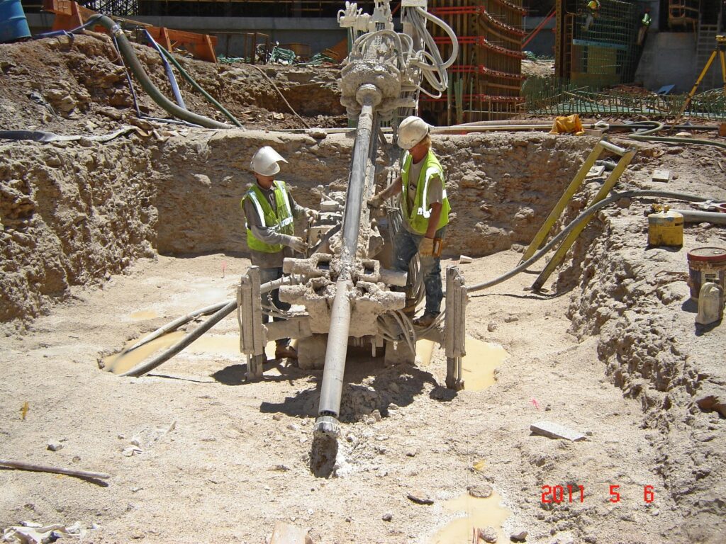 Two construction workers in safety gear operate drilling equipment at a construction site. The machinery is positioned in a muddy pit, surrounded by various cables and tools. The scene takes place outdoors on a sunny day.