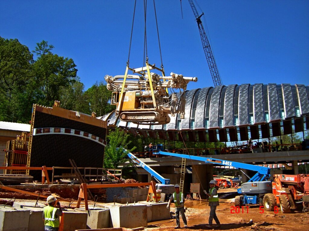 A large piece of machinery is being lifted by a crane at a construction site. Workers in safety gear observe the process. A partially constructed building and trees are visible in the background under a clear blue sky.