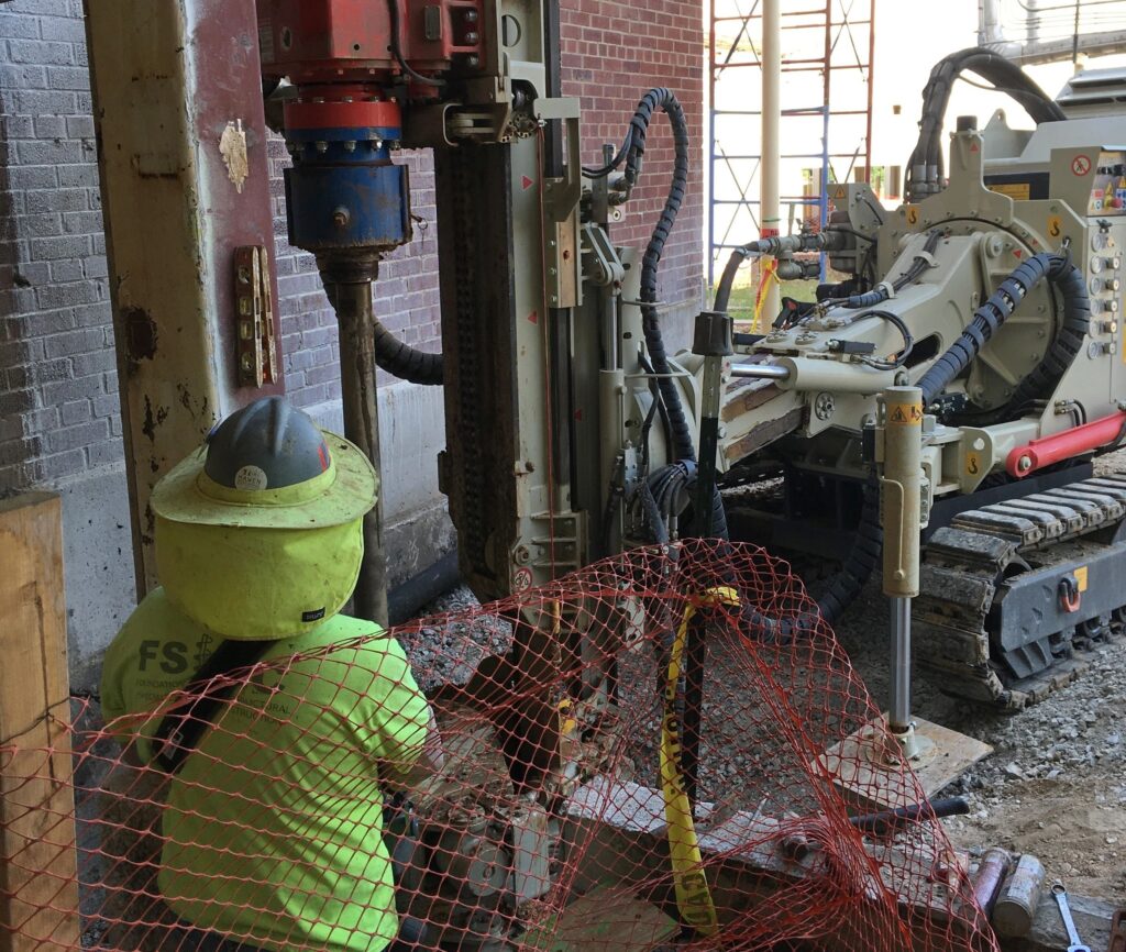 A construction worker wearing a helmet and yellow safety gear operates a large drilling machine inside a building under construction. The area is surrounded by safety netting, and a crawler machine is visible in the background.