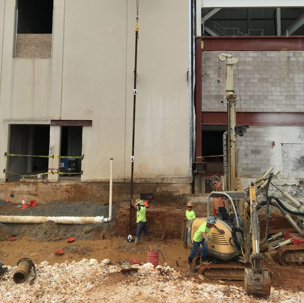 Construction workers in safety gear are operating machinery and installing a pipe at a building site. The area is muddy, with exposed concrete and metal beams visible on the unfinished structure.