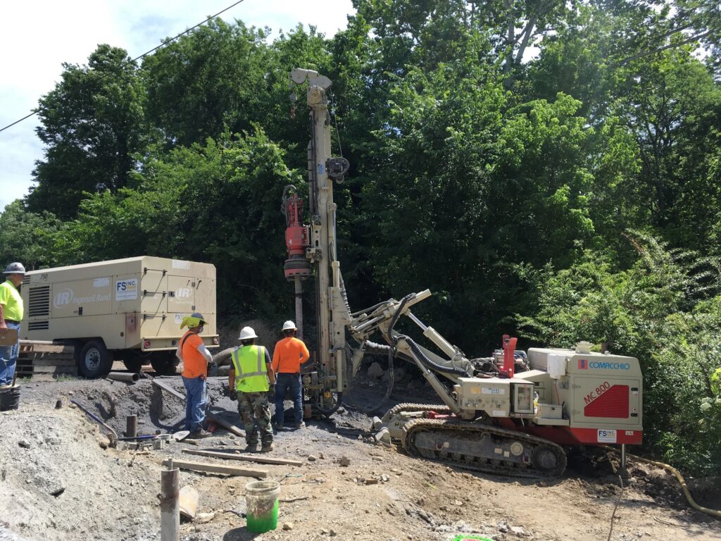 Construction workers operate a drilling machine on a roadside. They wear safety gear, including helmets and high-visibility vests. Trees and a dense green forest are in the background.