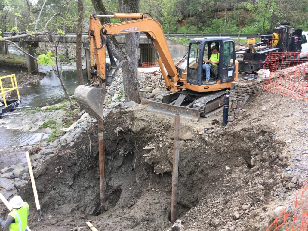 A construction site by a river with an excavator digging into the ground. Workers wearing safety vests and helmets are overseeing the operation. There are metal beams being installed and safety barriers surrounding the area.