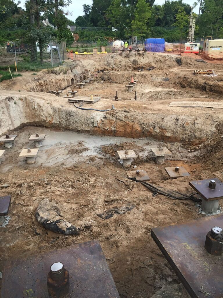 A construction site with deep excavated trenches and visible foundation pins. The area is surrounded by orange safety barriers, and there are trees and construction equipment in the background.