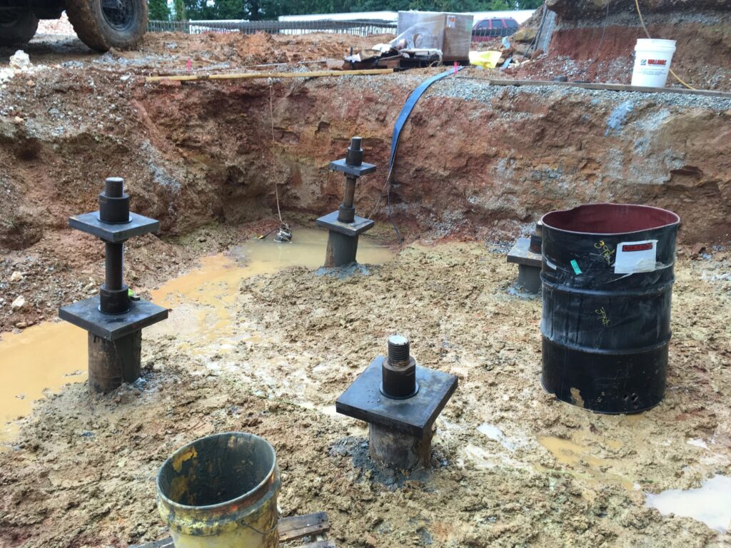 Construction site with metal foundation piers installed in a muddy pit. An overturned metal barrel and a plastic bucket are nearby. Soil and exposed earth surround the area. A construction vehicle is partially visible on the left.