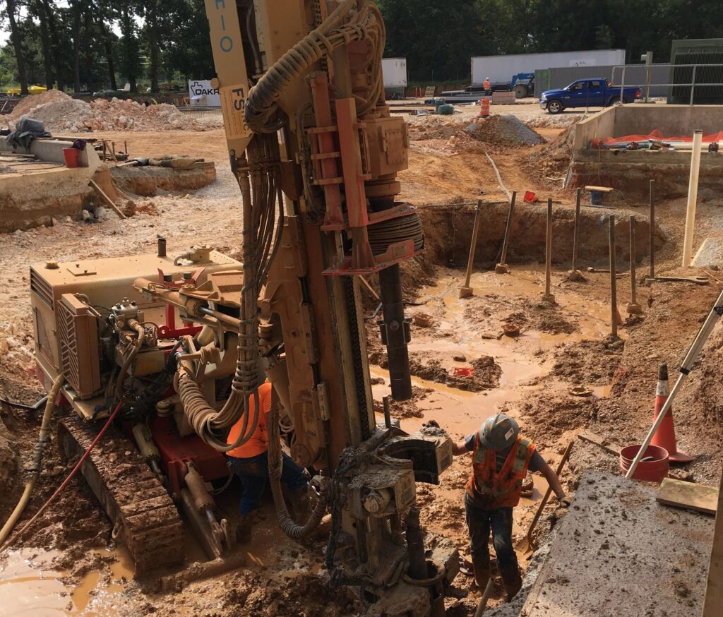 Construction workers drill at a muddy construction site with heavy machinery. Safety vests and helmets are worn. The area is surrounded by dirt and equipment, with trees and vehicles in the background.