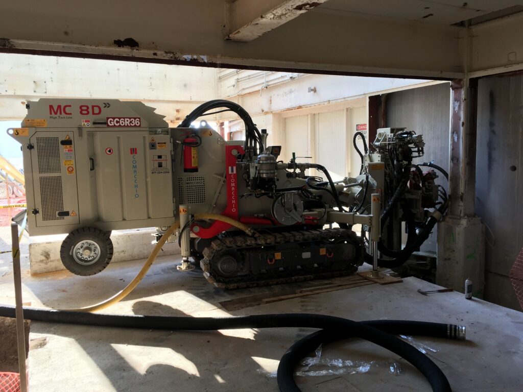 A drilling machine with crawler tracks and various hoses is set up inside a partially enclosed industrial space. Sunlight streams through open sections, highlighting the equipment's control panels and mechanical components.