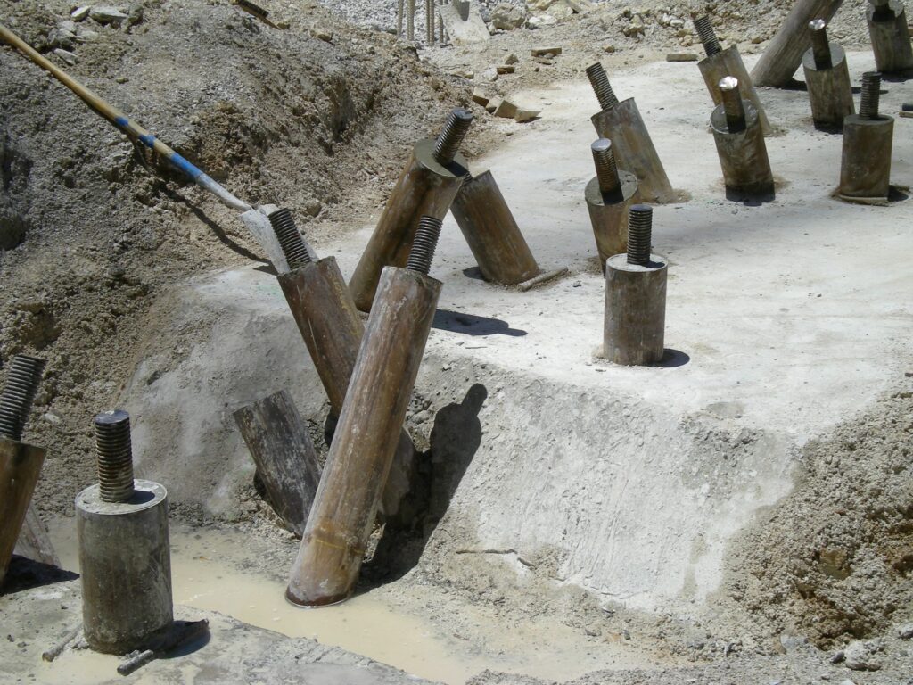 Construction site with several upright and tilted cylindrical steel beams embedded in concrete, surrounded by soil. The beams' tops have threaded sections, suggesting they are for structural support. The area appears to be under development.