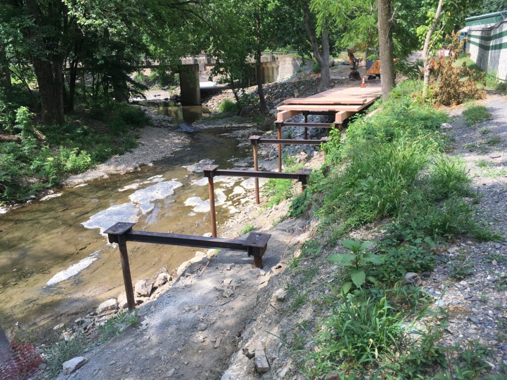 A construction site by a creek with unfinished bridge beams extending over a partially built concrete platform. Lush green trees and shrubs surround the area, and a small stream flows beneath the structure.