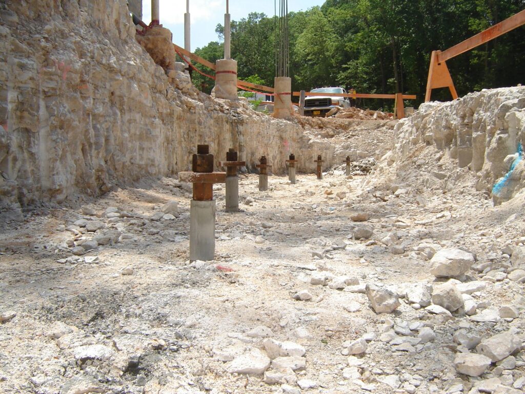 A construction site with exposed rocky terrain and steel foundation piles emerging from the ground. Wooden supports are visible in the background near trees and a parked vehicle. The sky is partly cloudy.